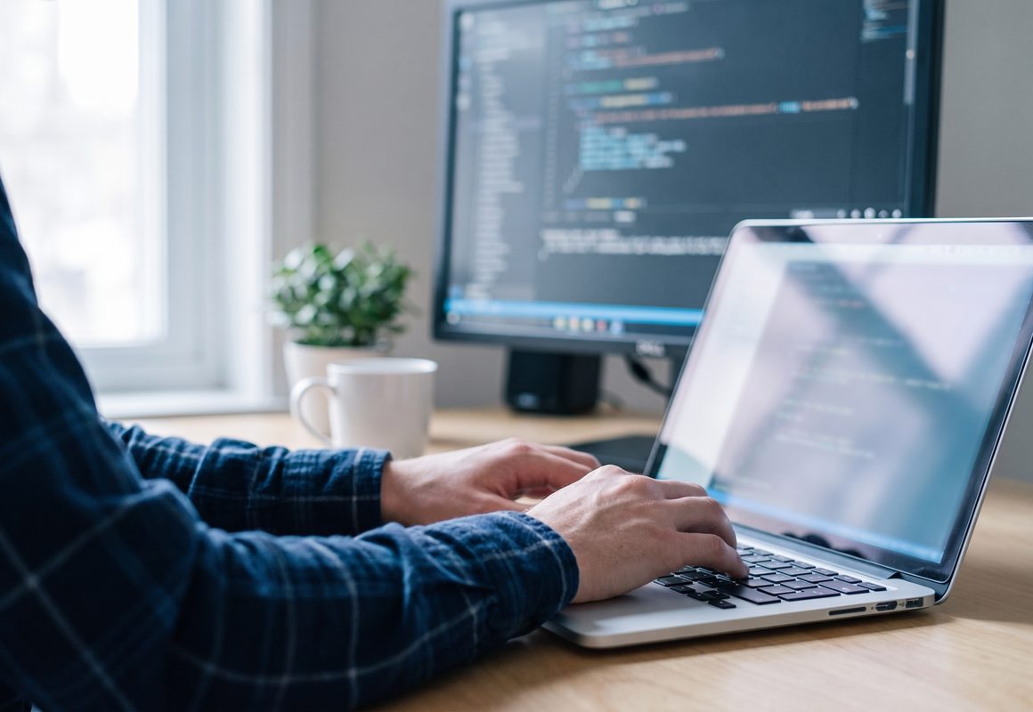 A developer typing on a laptop in front of a larger monitor, both showing source code